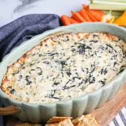 Spinach Artichoke Dip on wood board with crackers and vegetables.