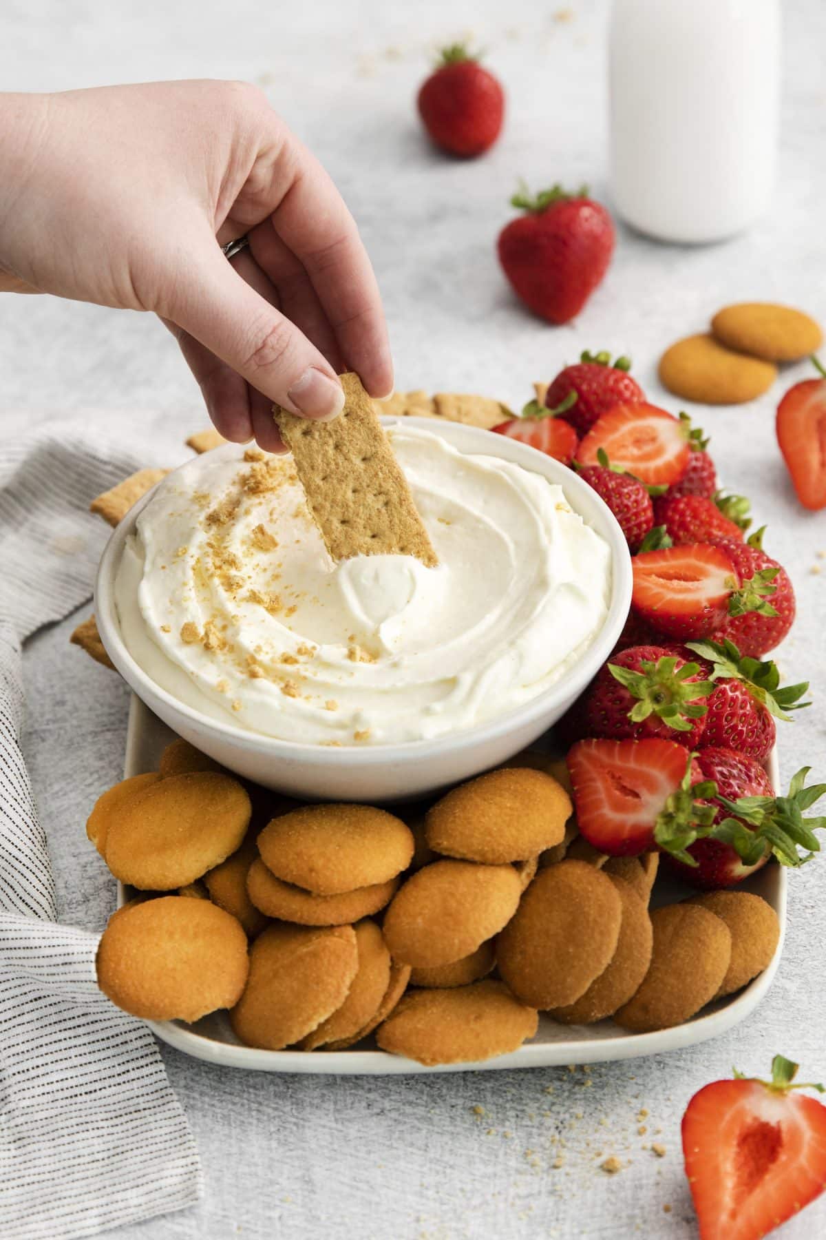 cheesecake dip in white bowl with hand, graham crackers, and strawberries.