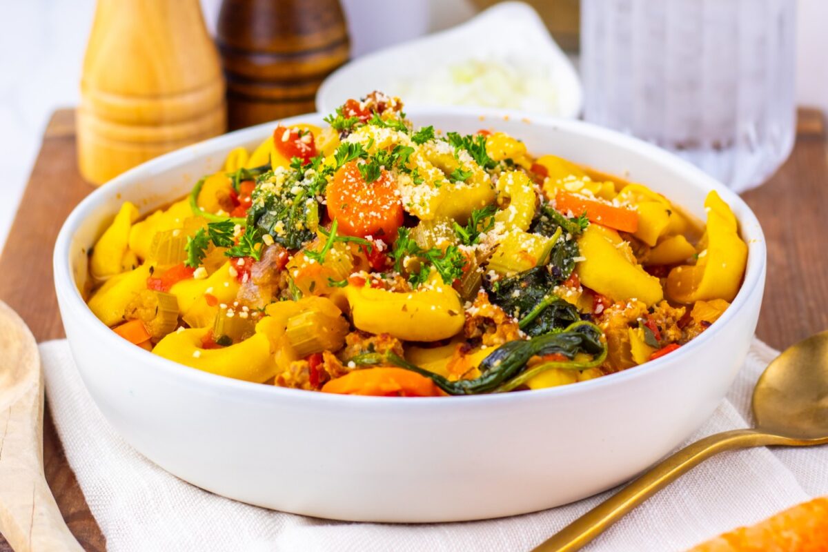 A bowl of sausage and butternut squash tortellini soup topped with shredded cheese and herbs.